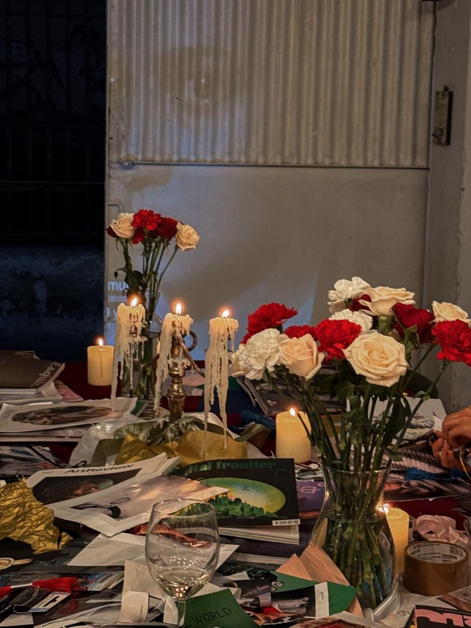 Candles burnt down over a table of open magazines and torn paper, the lit wall behind them.