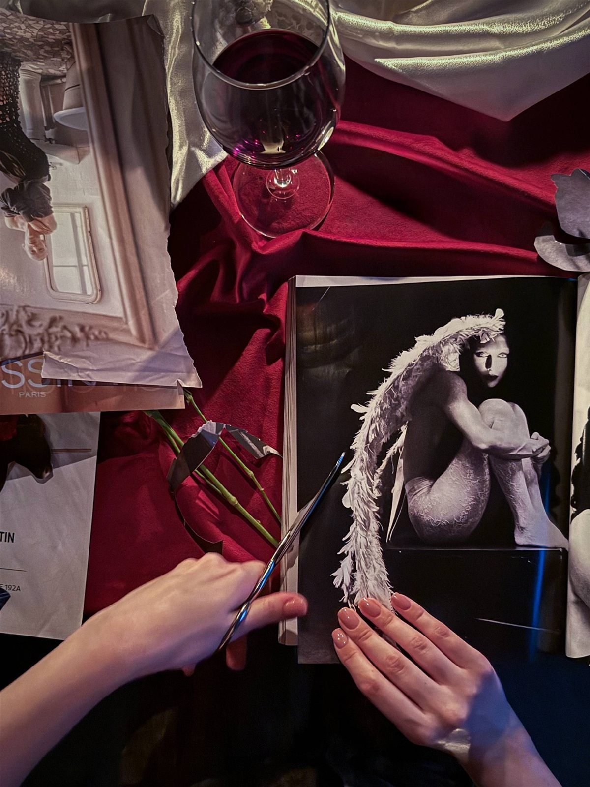 Two hands cutting a magazine spread with scissors on red velvet, beside a glass of red wine.